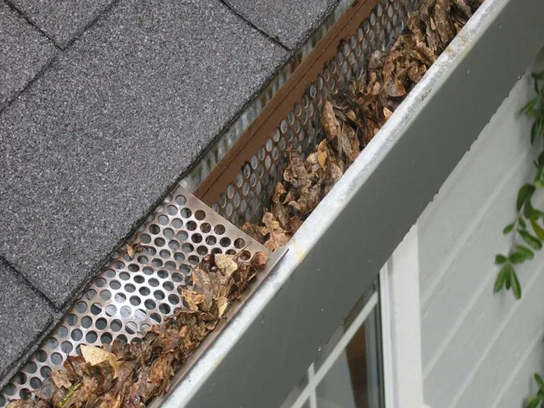 A perforated metal guard with leaves packed in the gutter beneath it